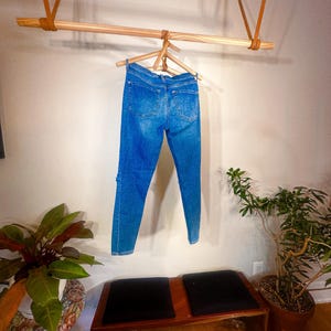 Op de afbeelding: Blauwe spijkerbroek hangt aan een houten kledingrek dat met leren riemen aan het plafond is bevestigd. De jeans worden gepresenteerd tegen een neutrale muur, met een bank en potplanten op de voorgrond.