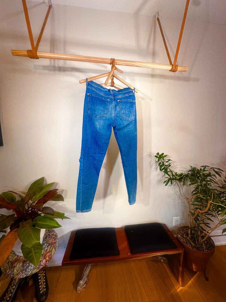 Op de afbeelding: Blauwe spijkerbroek hangt aan een houten kledingrek dat met leren riemen aan het plafond is bevestigd. De jeans worden gepresenteerd tegen een neutrale muur, met een bank en potplanten op de voorgrond.