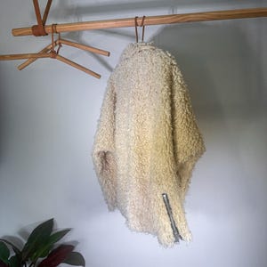 Puede incluir: Chaqueta texturizada de color crema con cremallera plateada. La chaqueta est&aacute; colgada de una percha de madera, mostrando su aspecto esponjoso y c&aacute;lido. El dise&ntilde;o sugiere una prenda de abrigo acogedora.