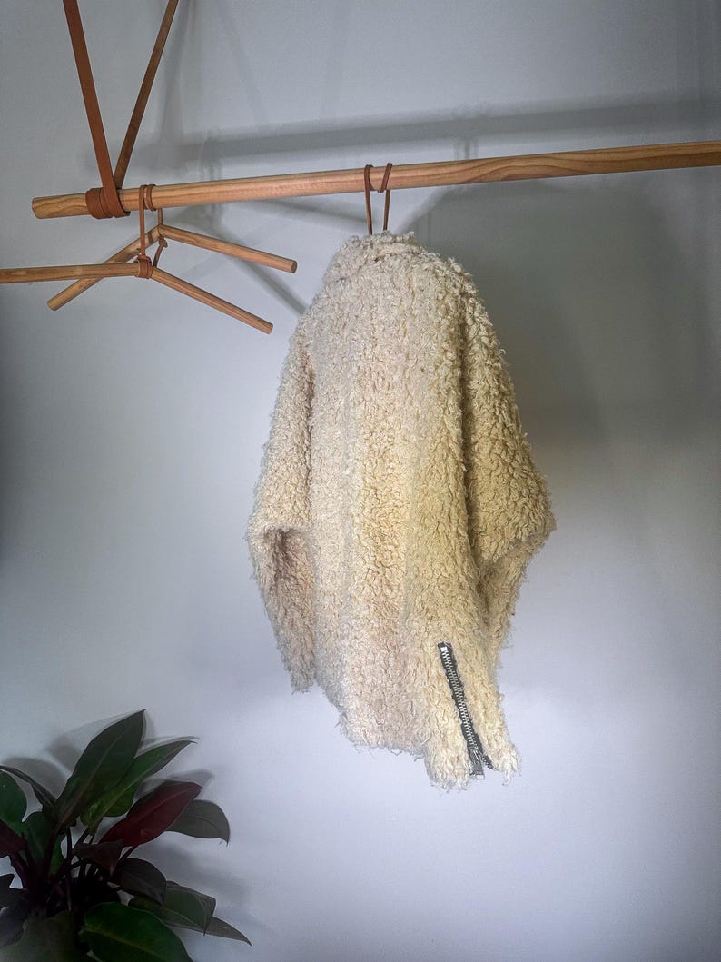 Puede incluir: Chaqueta texturizada de color crema con cremallera plateada. La chaqueta est&aacute; colgada de una percha de madera, mostrando su aspecto esponjoso y c&aacute;lido. El dise&ntilde;o sugiere una prenda de abrigo acogedora.