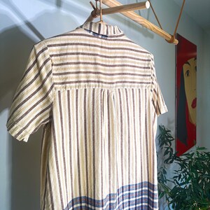 Op de afbeelding: Een gestreepte en geruite shirtjurk die aan een houten hanger hangt. De jurk heeft bruine en beige strepen met een blauw en bruin geruit patroon aan de onderkant.