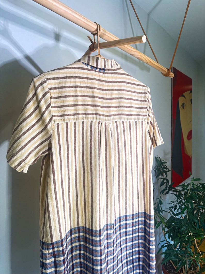 Op de afbeelding: Een gestreepte en geruite shirtjurk die aan een houten hanger hangt. De jurk heeft bruine en beige strepen met een blauw en bruin geruit patroon aan de onderkant.