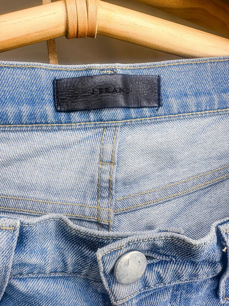 May include: Close-up of light blue denim jeans. The jeans feature a black leather tag with the brand name "J BRAND". The jeans have a button closure and visible stitching.
