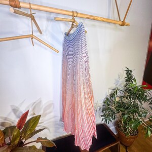 May include: A long, flowing skirt with a gradient pattern of blue, red, and white hangs on a wooden hanger. The skirt is displayed on a wooden rod suspended from the ceiling with leather straps. A bench and potted plants are in the foreground.