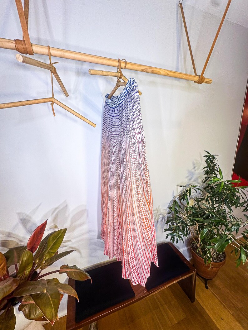 May include: A long, flowing skirt with a gradient pattern of blue, red, and white hangs on a wooden hanger. The skirt is displayed on a wooden rod suspended from the ceiling with leather straps. A bench and potted plants are in the foreground.