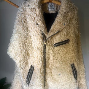 Puede incluir: Chaqueta de piel sint&eacute;tica color crema con un aspecto texturizado y rizado. La chaqueta presenta un estilo motero cl&aacute;sico con cuello de solapa, cierre de cremallera asim&eacute;trico y bolsillos con cremallera. La chaqueta est&aacute; colgada en una percha de madera.