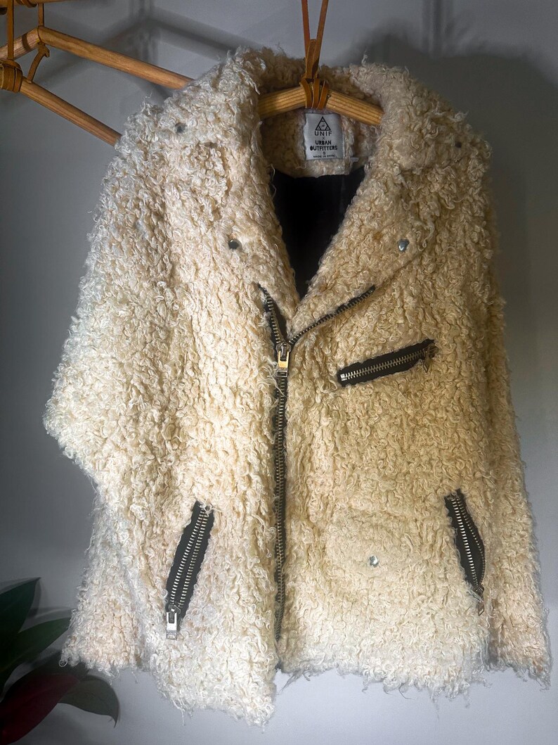 Puede incluir: Chaqueta de piel sint&eacute;tica color crema con un aspecto texturizado y rizado. La chaqueta presenta un estilo motero cl&aacute;sico con cuello de solapa, cierre de cremallera asim&eacute;trico y bolsillos con cremallera. La chaqueta est&aacute; colgada en una percha de madera.