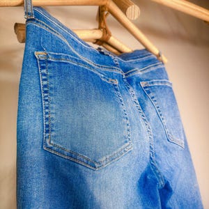 Op de afbeelding: Close-up van een blauwe spijkerbroek die aan een houten rek hangt. De jeans heeft zichtbare achterzakken en een klassiek vijf-zakken ontwerp. De denim heeft een vervaagde wassing.