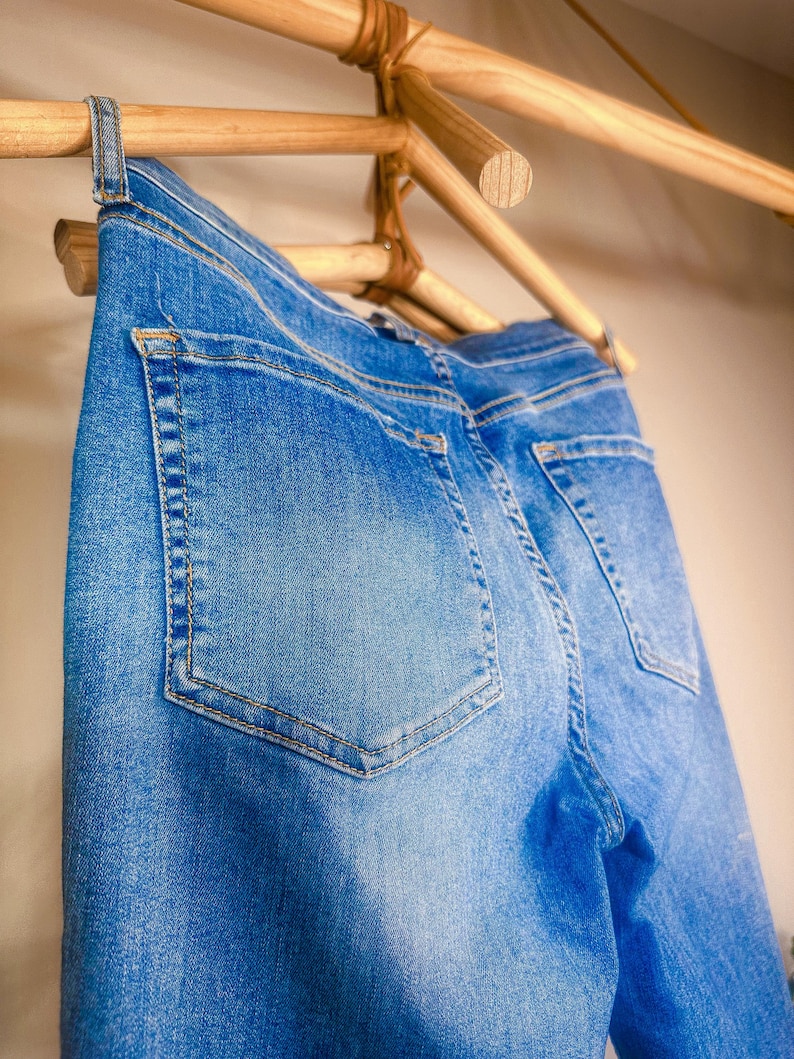 Op de afbeelding: Close-up van een blauwe spijkerbroek die aan een houten rek hangt. De jeans heeft zichtbare achterzakken en een klassiek vijf-zakken ontwerp. De denim heeft een vervaagde wassing.