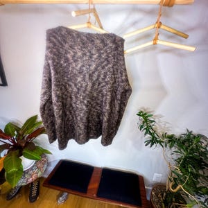 May include: A brown and beige fuzzy sweater hangs on a wooden clothes rack with leather straps. The rack is mounted to a white wall. A small wooden bench with black cushions sits in front of the rack.