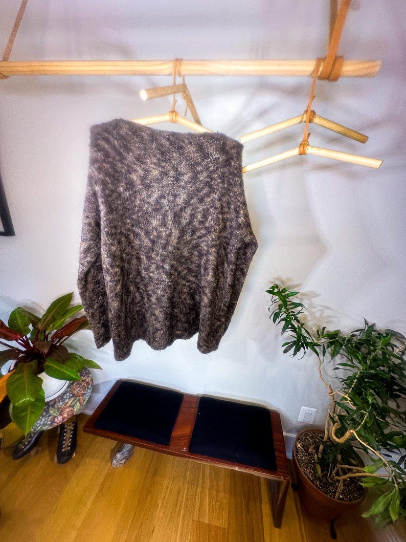 May include: A brown and beige fuzzy sweater hangs on a wooden clothes rack with leather straps. The rack is mounted to a white wall. A small wooden bench with black cushions sits in front of the rack.