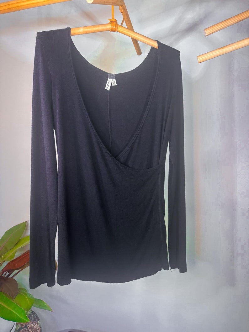 Puede incluir: Camiseta negra de manga larga con cuello en V y detalle de envoltura. La camiseta est&aacute; colgada en una percha de madera.
