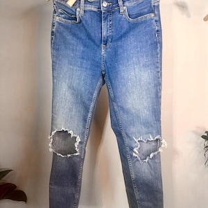 Op de afbeelding: Blauwe denim jeans met distressed knie-uitsparingen, hangend aan een houten hanger. De jeans hebben een klassiek ontwerp met vijf zakken en een vervaagde wassing. De jeans zijn van medium wash denim.