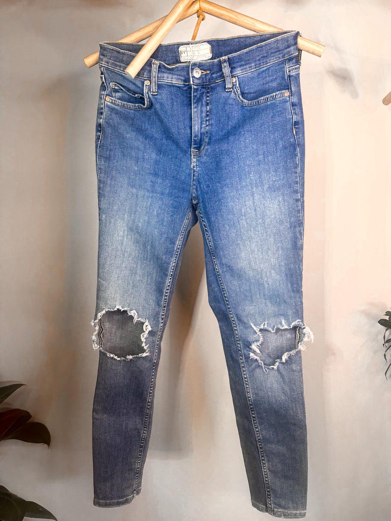 Op de afbeelding: Blauwe denim jeans met distressed knie-uitsparingen, hangend aan een houten hanger. De jeans hebben een klassiek ontwerp met vijf zakken en een vervaagde wassing. De jeans zijn van medium wash denim.