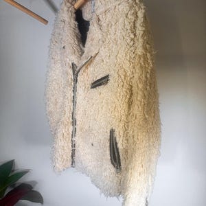 Puede incluir: Chaqueta texturizada de color crema con cremallera frontal y dos bolsillos con cremallera. La chaqueta est&aacute; colgada de una percha de madera. El material de la chaqueta tiene un aspecto lanudo y rizado.