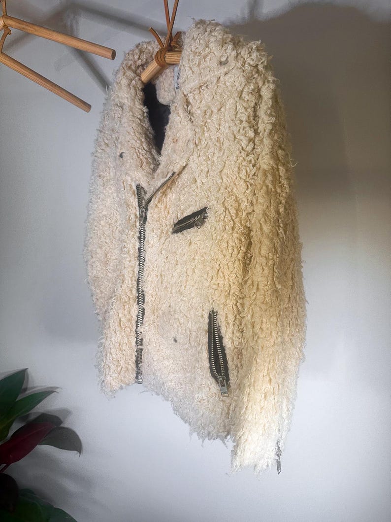 Puede incluir: Chaqueta texturizada de color crema con cremallera frontal y dos bolsillos con cremallera. La chaqueta est&aacute; colgada de una percha de madera. El material de la chaqueta tiene un aspecto lanudo y rizado.