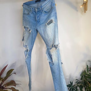 May include: Light blue denim jeans with distressed detailing, including large rips on the legs. The jeans are hanging on a wooden hanger, showcasing a casual, worn aesthetic. The jeans have a button fly and classic five-pocket design.