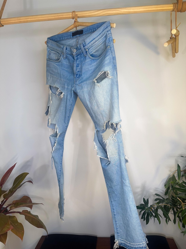 May include: Light blue denim jeans with distressed detailing, including large rips on the legs. The jeans are hanging on a wooden hanger, showcasing a casual, worn aesthetic. The jeans have a button fly and classic five-pocket design.