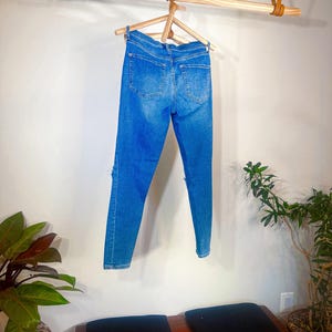 Op de afbeelding: Blauwe denim jeans met distressed details, hangend aan een houten kleerhanger. De jeans worden gepresenteerd tegen een witte muur, met een houten stang en leren riemen erboven. Een bank met zwarte kussens en potplanten staat eronder.