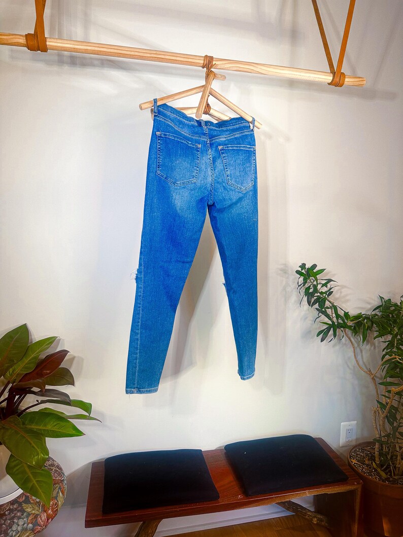 Op de afbeelding: Blauwe denim jeans met distressed details, hangend aan een houten kleerhanger. De jeans worden gepresenteerd tegen een witte muur, met een houten stang en leren riemen erboven. Een bank met zwarte kussens en potplanten staat eronder.