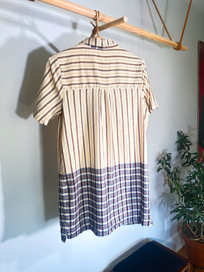 Op de afbeelding: Een shirt met korte mouwen met een gestreept patroon in bruin en beige. De onderste helft van het shirt is een ruitpatroon in blauw, bruin en wit.