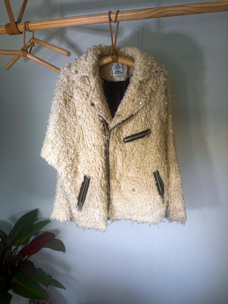 Puede incluir: Chaqueta crema texturizada con cierre de cremallera negro y dos bolsillos con cremallera. La chaqueta tiene un cuello ancho y est&aacute; colgada de una percha de madera. La chaqueta est&aacute; hecha de un material suave y difuso.