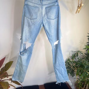 May include: Light blue denim jeans with distressed details, including rips on the knees and frayed hems. The jeans are displayed hanging on a wooden hanger, showcasing the back pockets and overall design. The jeans are a light wash.