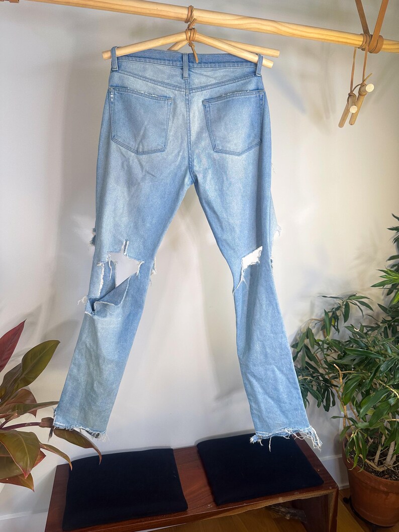 May include: Light blue denim jeans with distressed details, including rips on the knees and frayed hems. The jeans are displayed hanging on a wooden hanger, showcasing the back pockets and overall design. The jeans are a light wash.