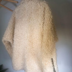Puede incluir: Chaqueta texturizada de color crema con un detalle de cremallera en la manga. La chaqueta est&aacute; colgada de una percha de madera. El material parece ser una tela suave y difusa.