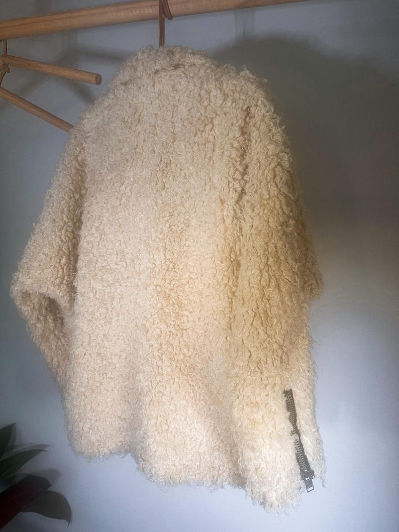 Puede incluir: Chaqueta texturizada de color crema con un detalle de cremallera en la manga. La chaqueta est&aacute; colgada de una percha de madera. El material parece ser una tela suave y difusa.