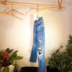 Op de afbeelding: Een paar blauwe spijkerbroeken met distressed details hangend aan een houten kledingrek.