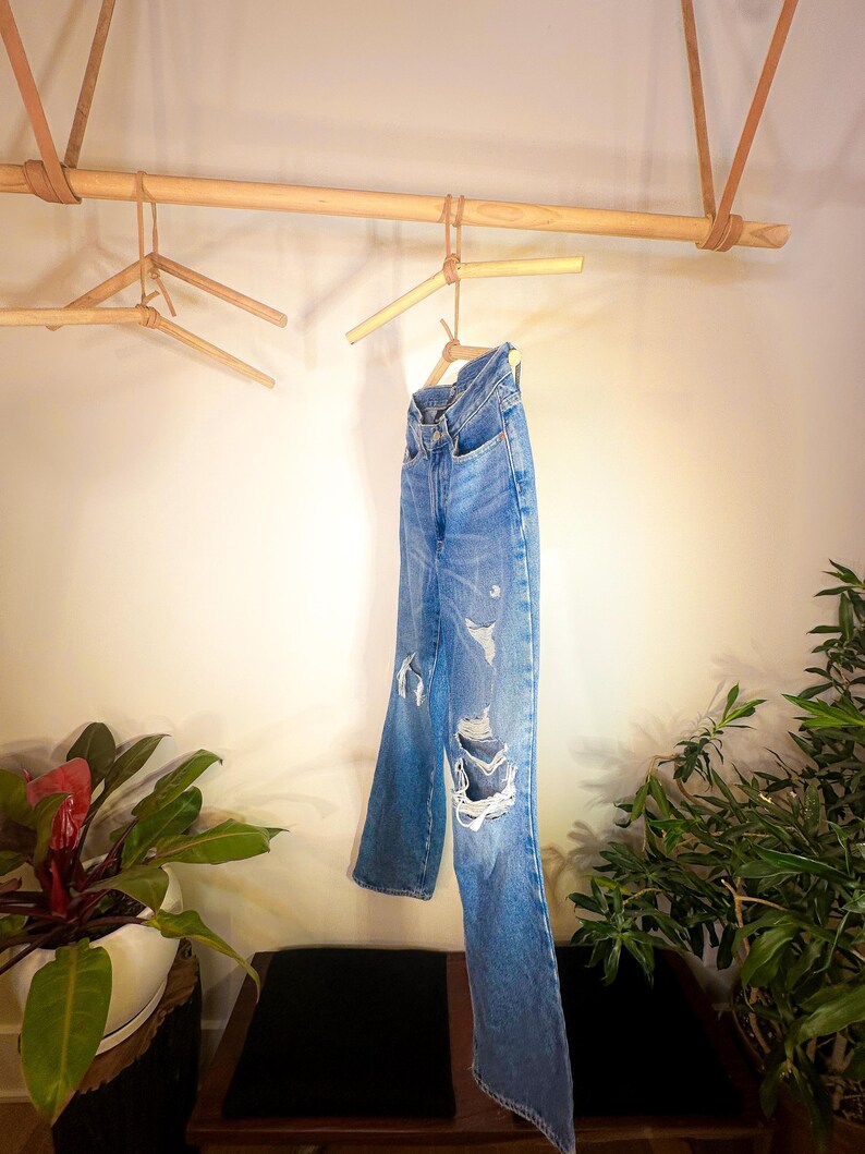Op de afbeelding: Een paar blauwe spijkerbroeken met distressed details hangend aan een houten kledingrek.