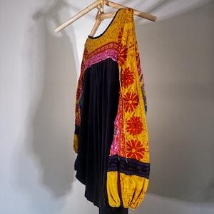 Puede incluir: Una camisa negra de manga larga con bordados florales amarillos, rosas y rojos en las mangas. La camisa est&aacute; colgada en una percha de madera.