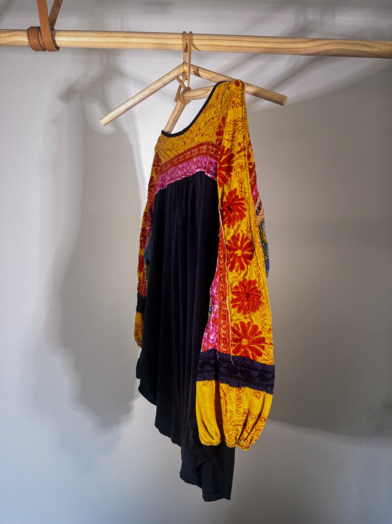 Puede incluir: Una camisa negra de manga larga con bordados florales amarillos, rosas y rojos en las mangas. La camisa est&aacute; colgada en una percha de madera.