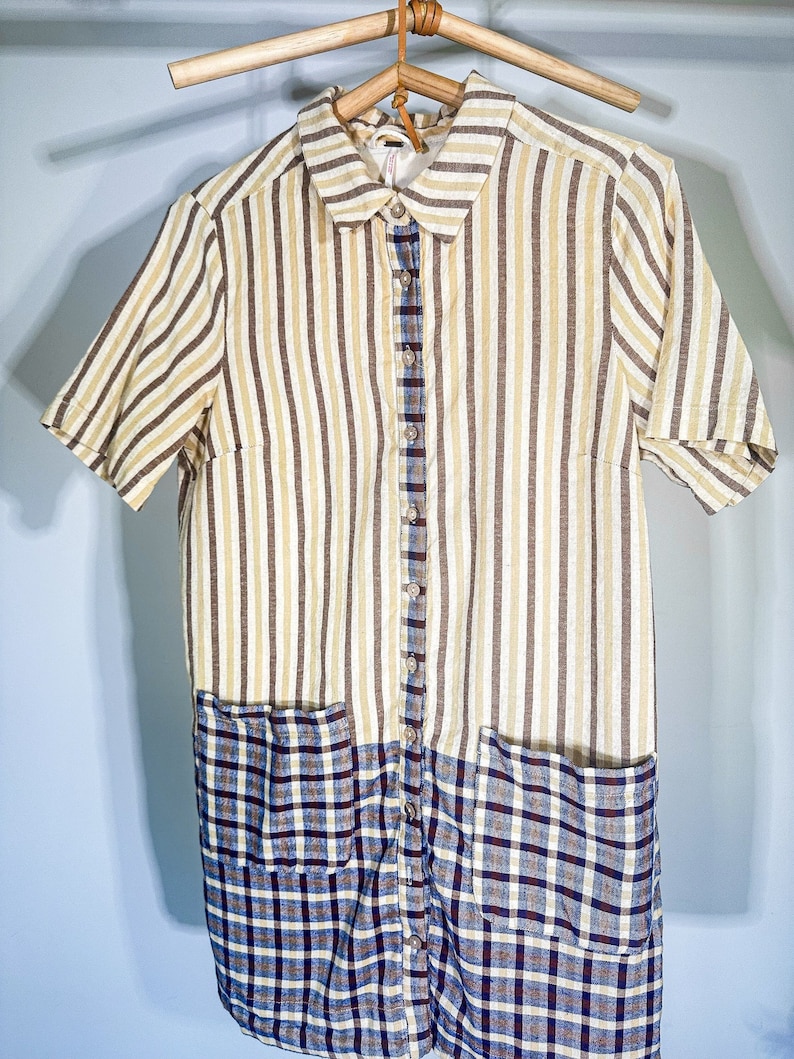 May include: A striped and plaid shirt dress with short sleeves and two pockets. The dress is a light yellow and brown striped fabric with a blue and brown plaid fabric on the bottom half. The dress has a button-up front and a collared neckline.
