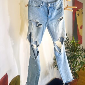 May include: Light blue denim jeans with distressed detailing, featuring large rips on the legs. The jeans are suspended from a wooden rack, highlighting their design. They have a button closure and classic five-pocket styling.