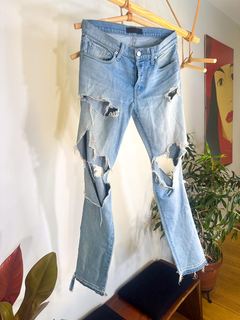 May include: Light blue denim jeans with distressed detailing, featuring large rips on the legs. The jeans are suspended from a wooden rack, highlighting their design. They have a button closure and classic five-pocket styling.