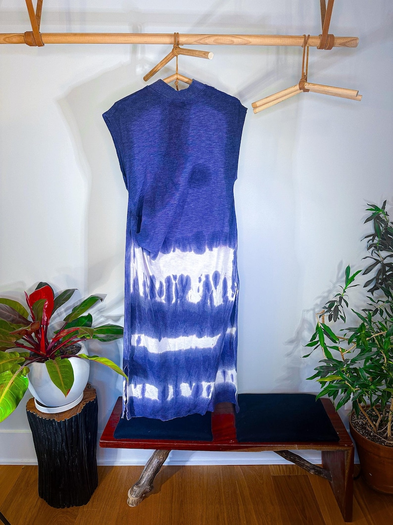 May include: A blue and white tie-dye sleeveless maxi dress hanging on a wooden hanger. The dress is hanging in a room with a wooden bench and plants.
