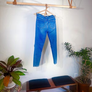 Op de afbeelding: Blauwe spijkerbroek hangend aan een houten hanger. De jeans worden gepresenteerd tegen een witte muur, met een houten stang en leren riemen erboven. Een bank met zwarte kussens en potplanten staan op de voorgrond.