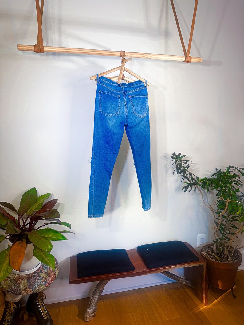 Op de afbeelding: Blauwe spijkerbroek hangend aan een houten hanger. De jeans worden gepresenteerd tegen een witte muur, met een houten stang en leren riemen erboven. Een bank met zwarte kussens en potplanten staan op de voorgrond.