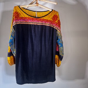 Puede incluir: Una camisa negra de manga larga con un cuello y mangas con estampado amarillo, rojo y azul. La camisa est&aacute; colgada de una percha de madera.