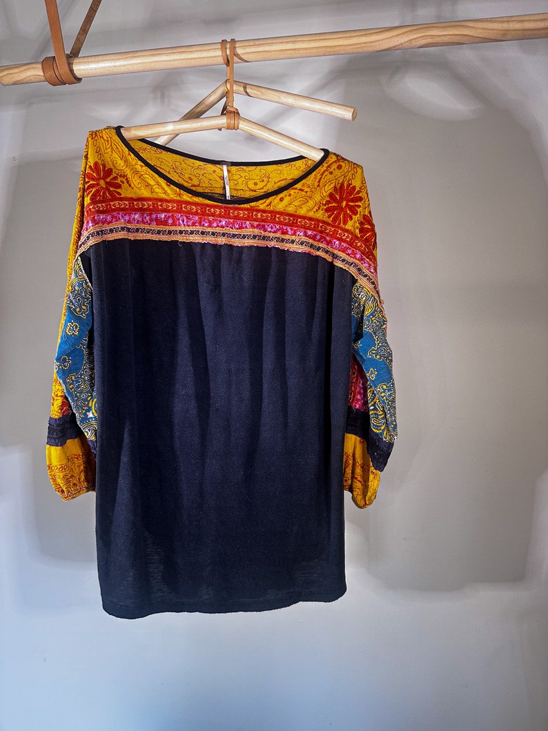 Puede incluir: Una camisa negra de manga larga con un cuello y mangas con estampado amarillo, rojo y azul. La camisa est&aacute; colgada de una percha de madera.