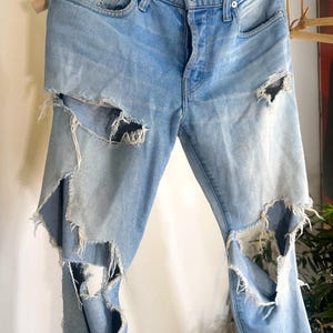May include: Light blue denim jeans with distressed detailing. The jeans have multiple large rips on the front of the legs, with frayed edges. They feature a button and zip closure, and classic five-pocket styling.