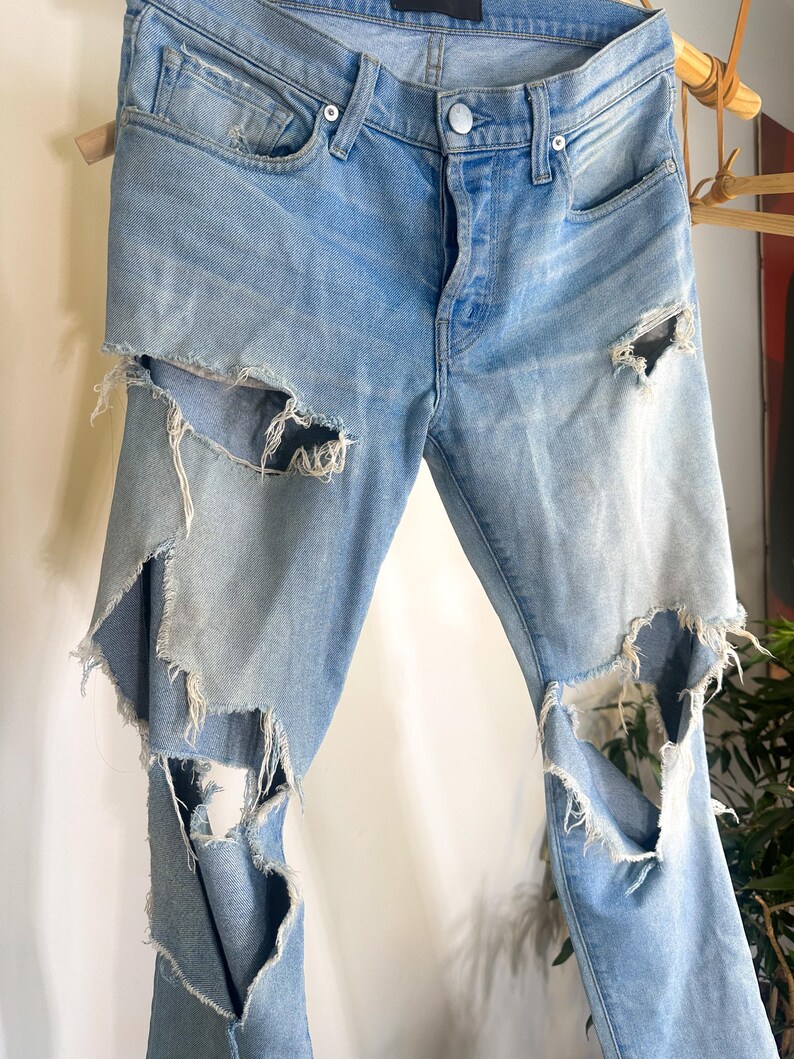 May include: Light blue denim jeans with distressed detailing. The jeans have multiple large rips on the front of the legs, with frayed edges. They feature a button and zip closure, and classic five-pocket styling.