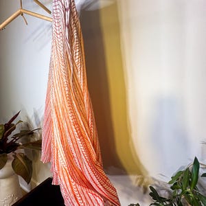 May include: A long, flowing fabric with a gradient pattern of red, orange, and white stripes. The fabric is hanging from a wooden rack, with the stripes creating a vertical design. The overall aesthetic is elegant and airy.