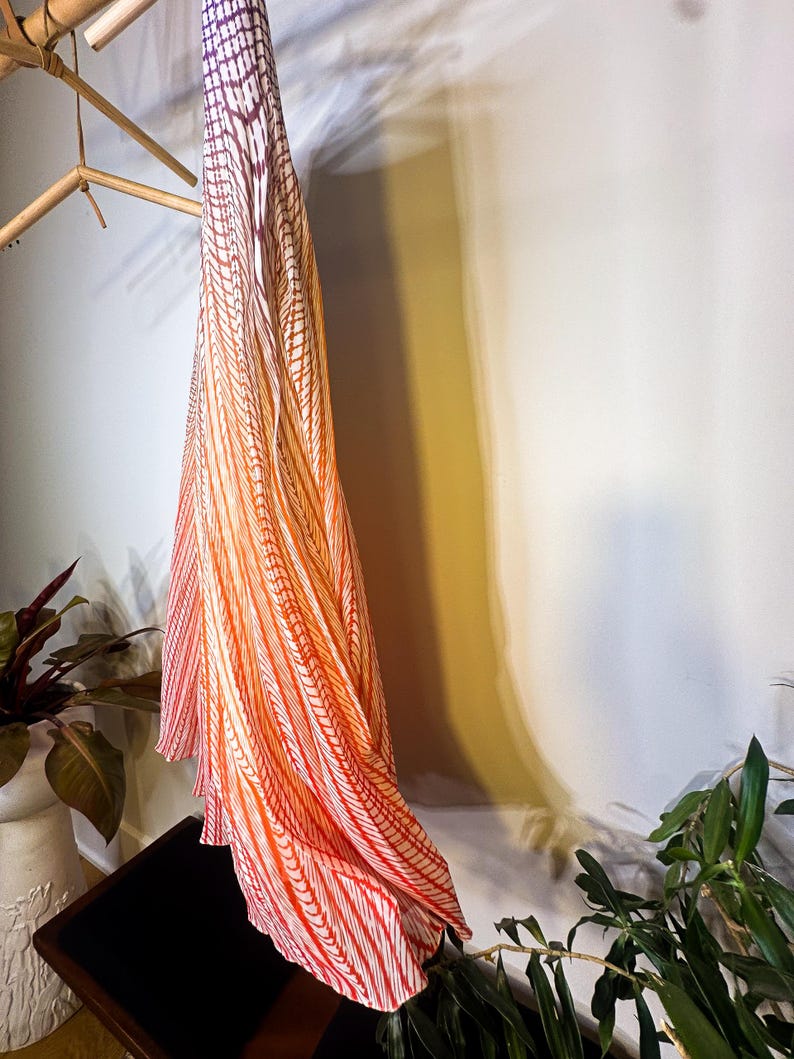 May include: A long, flowing fabric with a gradient pattern of red, orange, and white stripes. The fabric is hanging from a wooden rack, with the stripes creating a vertical design. The overall aesthetic is elegant and airy.