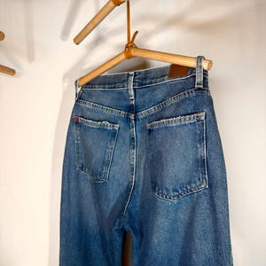 Op de afbeelding: Een paar blauwe denim jeans hangend aan een houten hanger. De jeans heeft een klassieke rechte pijp en is gemaakt van een zachte, comfortabele stof.