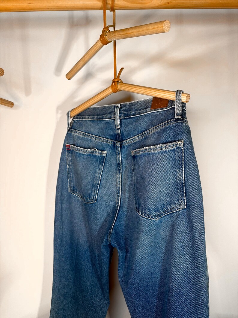 Op de afbeelding: Een paar blauwe denim jeans hangend aan een houten hanger. De jeans heeft een klassieke rechte pijp en is gemaakt van een zachte, comfortabele stof.