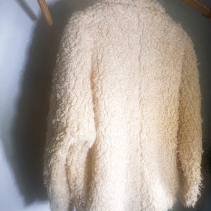 Puede incluir: Chaqueta crema texturizada colgada de una percha de madera. La chaqueta tiene una textura gruesa y rizada y un dise&ntilde;o cl&aacute;sico. El color neutro y la apariencia acogedora sugieren una prenda de abrigo c&aacute;lida y elegante.