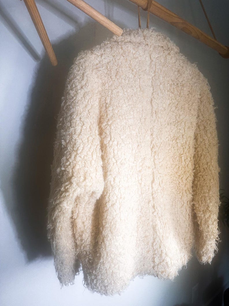 Puede incluir: Chaqueta crema texturizada colgada de una percha de madera. La chaqueta tiene una textura gruesa y rizada y un dise&ntilde;o cl&aacute;sico. El color neutro y la apariencia acogedora sugieren una prenda de abrigo c&aacute;lida y elegante.